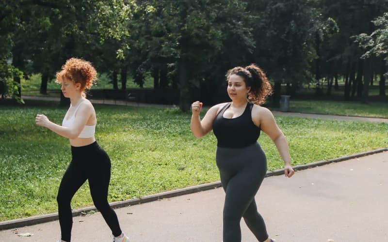 Duas mulheres caminham juntas por uma pista arborizada em um parque.