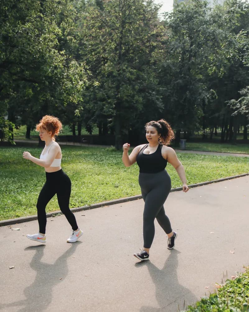Duas mulheres caminham juntas por uma pista arborizada em um parque.