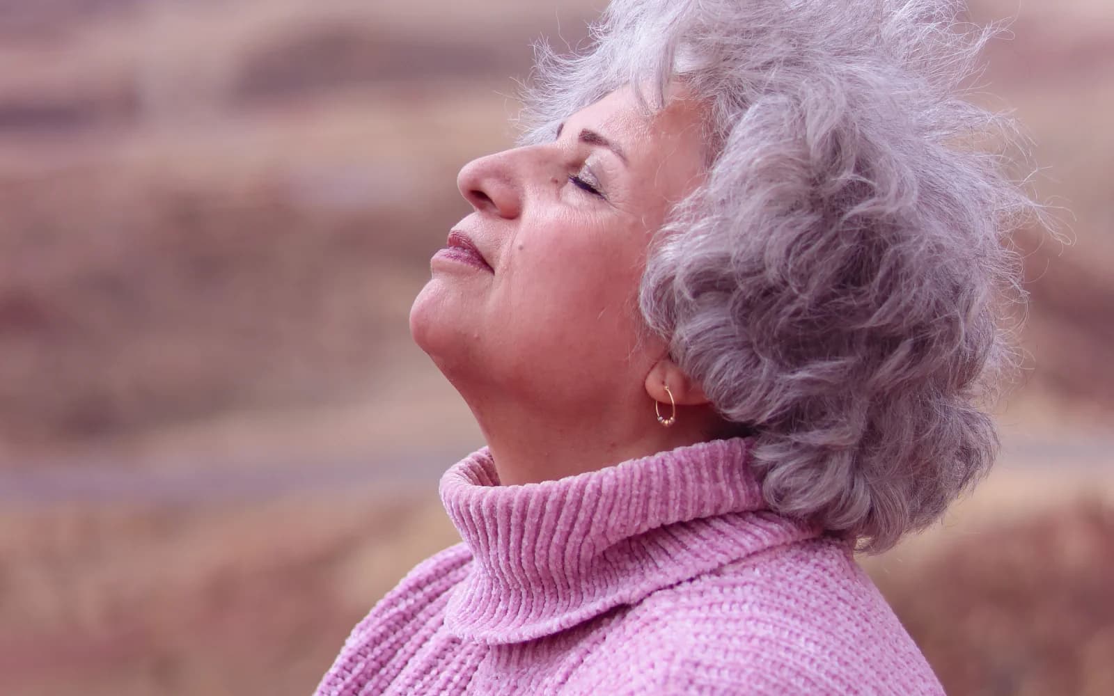 Mulher idosa de olhos fechados respira ao ar livre diante de uma paisagem natural.